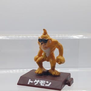 May include: A yellow plastic figurine of a creature wearing sunglasses, standing on a brown base with the Japanese text "トガモン".