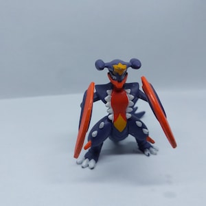 Puede incluir: Una figura de acción de Pokemon de color morado oscuro y rojo con alas naranjas. La figura tiene una estrella amarilla en la cabeza y dientes blancos.