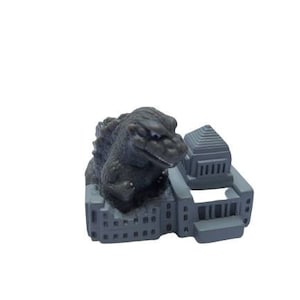 May include: A gray plastic toy of Godzilla destroying a building. The building is a simple model of a building with a flat roof and windows.