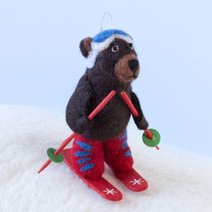 May include: A brown felt bear wearing a blue and white ski hat and red ski pants is skiing down a snowy hill. The bear is holding red ski poles with green circular bases.