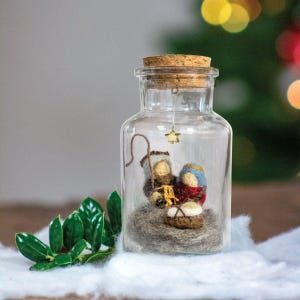 May include: A glass jar with a cork lid containing a felt nativity scene. The scene includes Mary, Joseph, and baby Jesus. A small gold star hangs from the top of the jar.