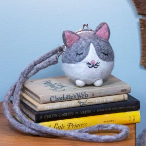 May include: A gray and white felt cat-shaped coin purse with a silver clasp and a gray strap. The cat has pink ears and closed eyes. The purse sits atop a stack of vintage books, including "Charlotte's Web."