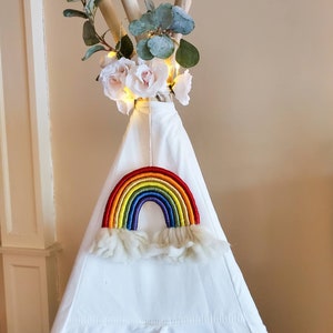 May include: A white teepee tent with a rainbow-colored yarn rainbow hanging from the top. The teepee is decorated with white flowers and greenery.
