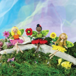 May include: Three felt fairy figurines with wings and flowers. The fairies are wearing colorful dresses and have different skin tones. The flowers are pink, red, and yellow.