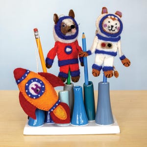 May include: A whimsical desk organizer featuring felt astronaut animal figures and a rocket ship. The figures are in red and white space suits. The rocket is orange and red. Pencils and pens are held in blue, gray, and green holders.