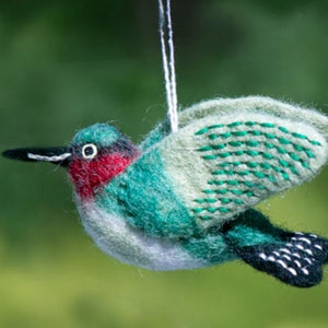 May include: A felt hummingbird ornament with green and white wings, a red throat, and black eyes. The ornament is hanging from a white string.