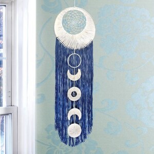May include: A handcrafted dream catcher with a white woven web and a blue tassel design. The dream catcher features a circular design with phases of the moon, including crescent and full moon shapes, hanging below.