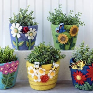 May include: Five felt flower pots with different floral designs and butterflies. The pots are blue, green, yellow, and turquoise. The pots are filled with small plants.