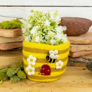 May include: A felted yellow and brown beehive-shaped planter with white daisy accents and a red door. A felted bee is attached to the side. The planter is filled with white flowers. The planter is sitting on a wooden surface.