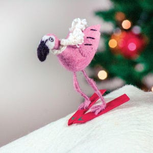 May include: A pink felt flamingo wearing a white felt hat and skiing on red skis. The flamingo is on a white surface with a blurred Christmas tree in the background.