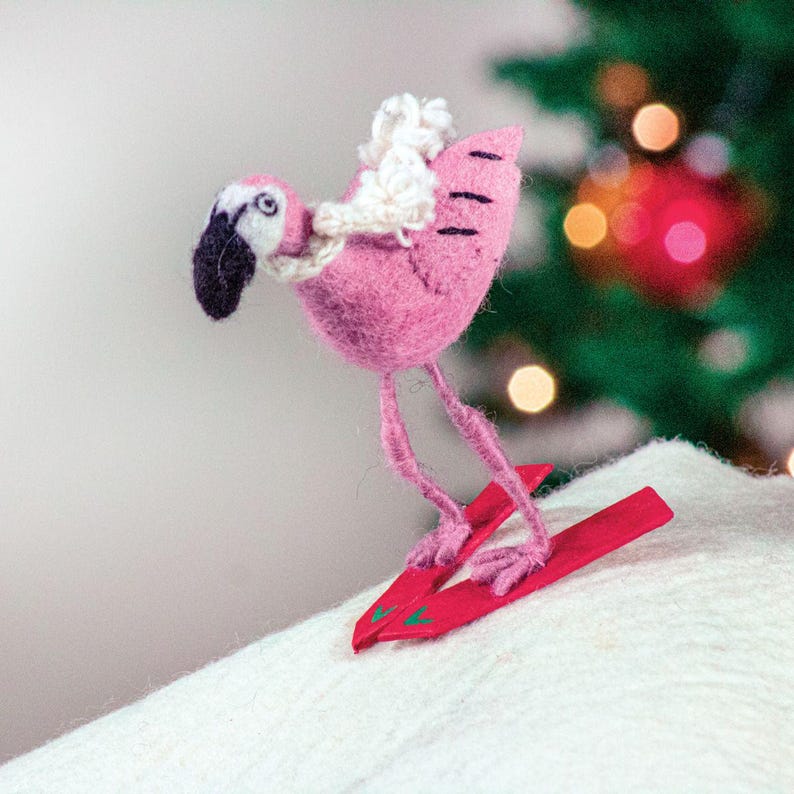 May include: A pink felt flamingo wearing a white felt hat and skiing on red skis. The flamingo is on a white surface with a blurred Christmas tree in the background.