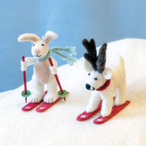 May include: Two felt animal figurines, a white rabbit and a reindeer, are skiing on red skis. The rabbit is wearing a blue scarf and the reindeer has a red scarf. The figurines are on a white surface.