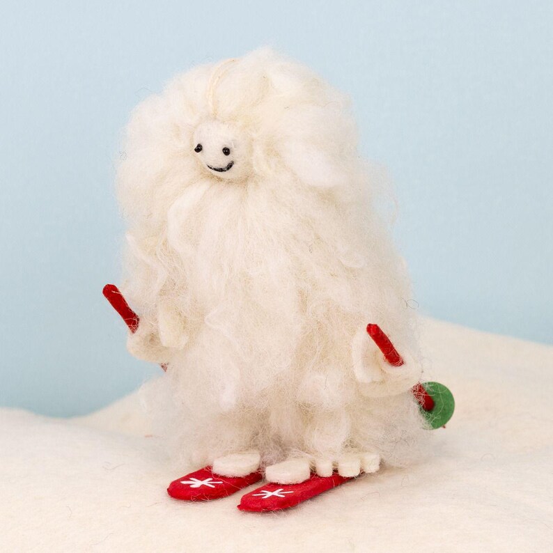 May include: A white felt snowman ornament with a smiling face, wearing red ski boots and holding red skis.