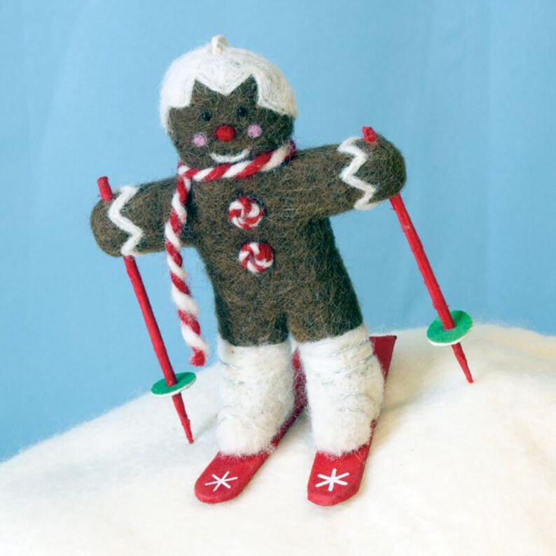May include: A gingerbread man wearing a white hat and red and white striped scarf is skiing down a snowy hill. He is holding two red ski poles with green rings at the ends. He is wearing white boots and red skis with white snowflakes on them.