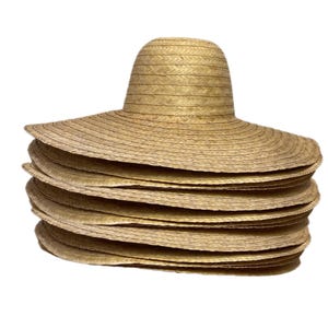 May include: A pile of wide-brimmed straw hats in a natural colour. Each hat has a rounded crown and a broad, circular brim. The hats are made of woven straw, and the stack shows the texture and layers of the hats.