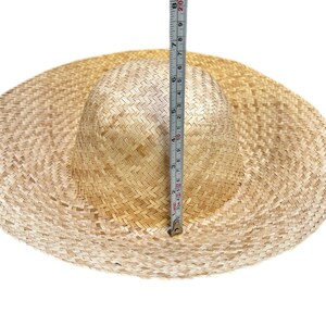 Puede incluir: Un sombrero de paja trenzada de color tostado claro con ala ancha y copa redondeada. Una cinta m&eacute;trica vertical est&aacute; colocada contra el centro de la copa, mostrando una altura de aproximadamente 20 cm desde la base hasta la parte superior.