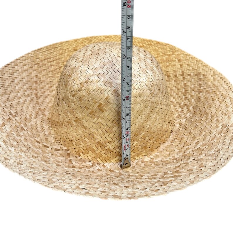 Puede incluir: Un sombrero de paja trenzada de color tostado claro con ala ancha y copa redondeada. Una cinta m&eacute;trica vertical est&aacute; colocada contra el centro de la copa, mostrando una altura de aproximadamente 20 cm desde la base hasta la parte superior.