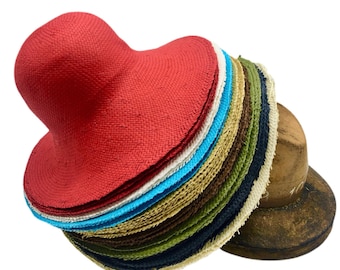 May include: A stack of woven straw hats in various colors including red, white, turquoise, tan, brown, green, and navy blue. The hats are layered on top of each other, with the bright red hat on the very top, resting against a wooden hat block form.