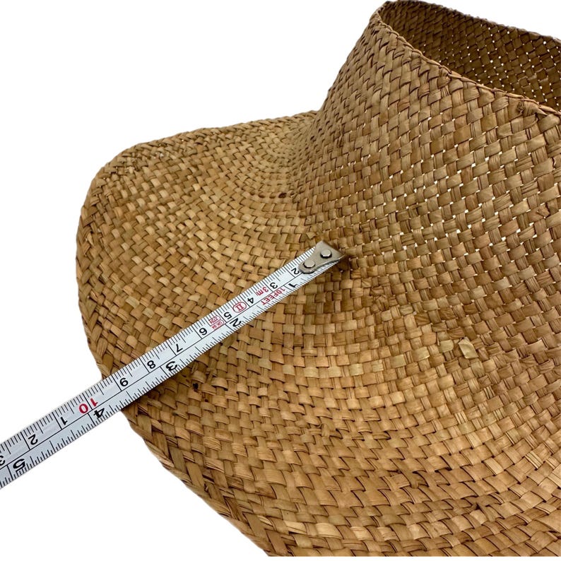 May include: A woven straw basket with a wide brim, measured with a tape measure. The basket is a natural tan colour and has a textured surface. The tape measure shows a measurement of approximately 25 cm.