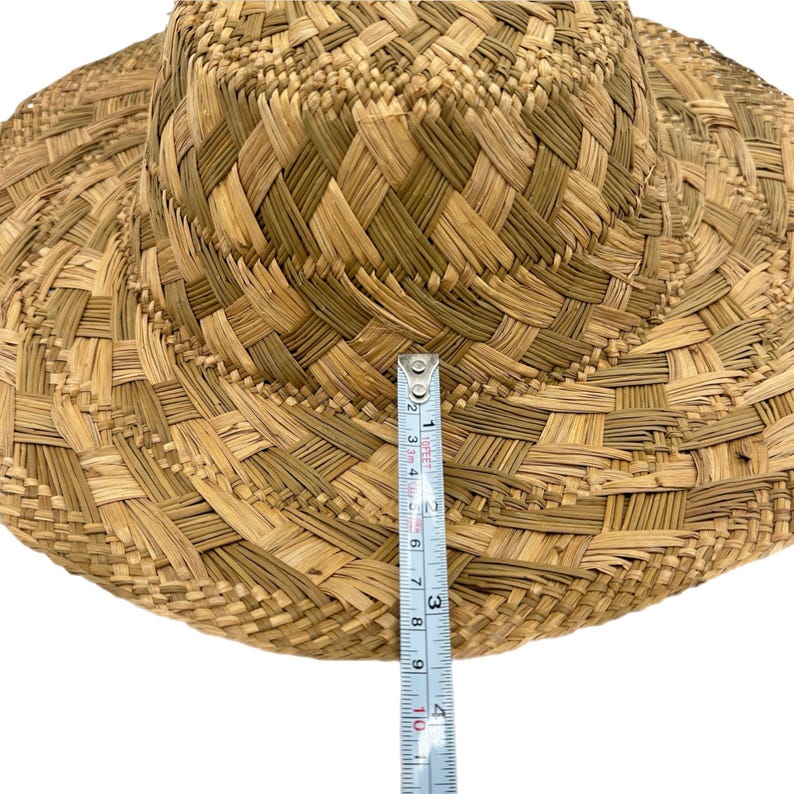 May include: A woven straw hat with a wide brim. The hat is tan and brown with a textured pattern. The brim measures approximately 25 cm in diameter. A measuring tape is placed on the hat.