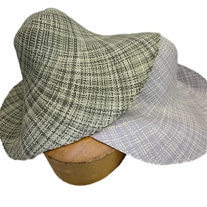 May include: Two woven sun hats in shades of green and lavender. The hats have wide brims and a textured, basket-weave pattern. They are displayed on a wooden hat form, showcasing their shape and design. The hats are made of a lightweight material.