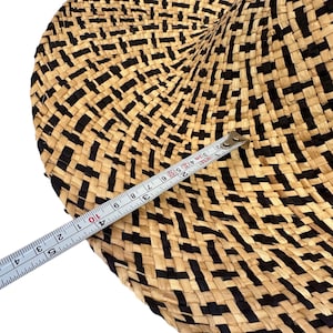 May include: A round woven item with a black and beige geometric design. A measuring tape indicates a measurement of approximately 25 cm. The item appears to be made from natural fibres.