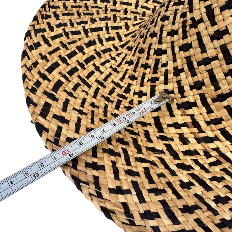 May include: A round woven item with a black and beige geometric design. A measuring tape indicates a measurement of approximately 25 cm. The item appears to be made from natural fibres.
