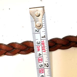 May include: Close-up of a brown leather braided belt measured with a tape measure. The tape measure reads 10 feet and 2 inches.