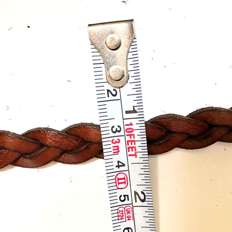 May include: Close-up of a brown leather braided belt measured with a tape measure. The tape measure reads 10 feet and 2 inches.