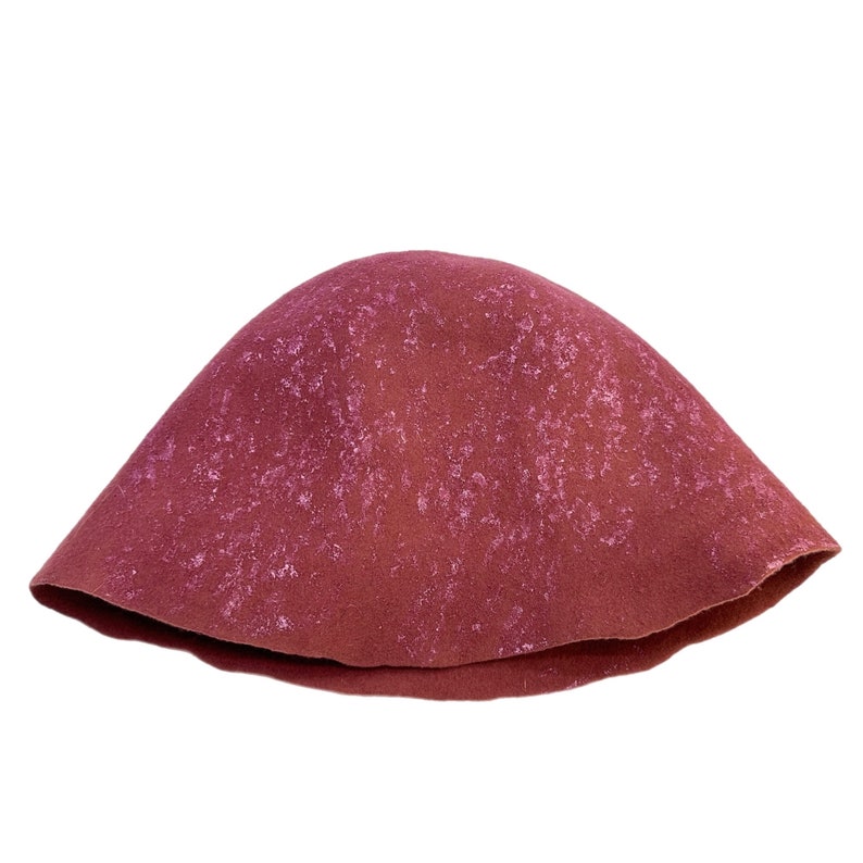 Może przedstawiać: Bordowy kapelusz typu cloche z teksturowaną powierzchnią. Kapelusz ma zaokrągloną koronę i lekko rozszerzane rondo. Kolor to głęboki czerwonobrązowy z jaśniejszymi, nakrapianymi akcentami.