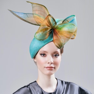 Op de afbeelding: Een turquoise hoed met een grote, decoratieve strik in tinten oranje, groen en blauw. Deze elegante hoofdbedekking is een opvallend accessoire, geschikt voor formele evenementen en speciale gelegenheden.