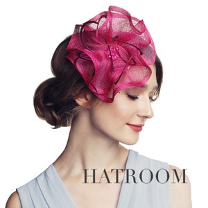 May include: A pink and white feathered hat with a large bow. The hat is worn by a woman with her hair pulled back.