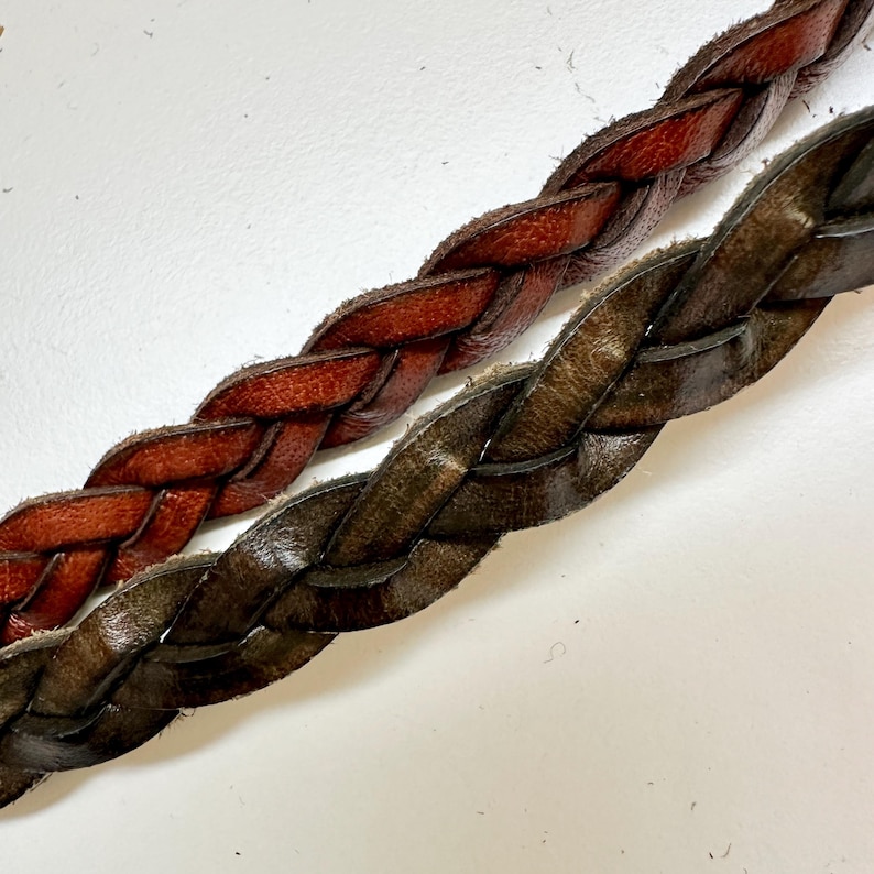 May include: Two braided leather straps, one brown and one dark green, are laid side by side. The straps are woven together in a simple braid pattern.
