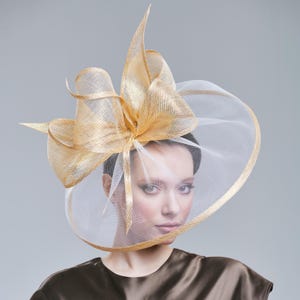 May include: A gold and white formal hat with a large bow and a wide brim. The hat has a sheer veil that covers the face. The hat is made of a woven material and has a metallic sheen. The hat is worn by a person.