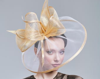 May include: A gold and white formal hat with a large bow and a wide brim. The hat has a sheer veil that covers the face. The hat is made of a woven material and has a metallic sheen. The hat is worn by a person.