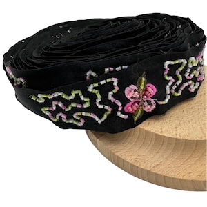 May include: A roll of black fabric ribbon with floral and geometric beaded embroidery. The embroidery features pink, green, and white beads. The ribbon is resting on a wooden surface.