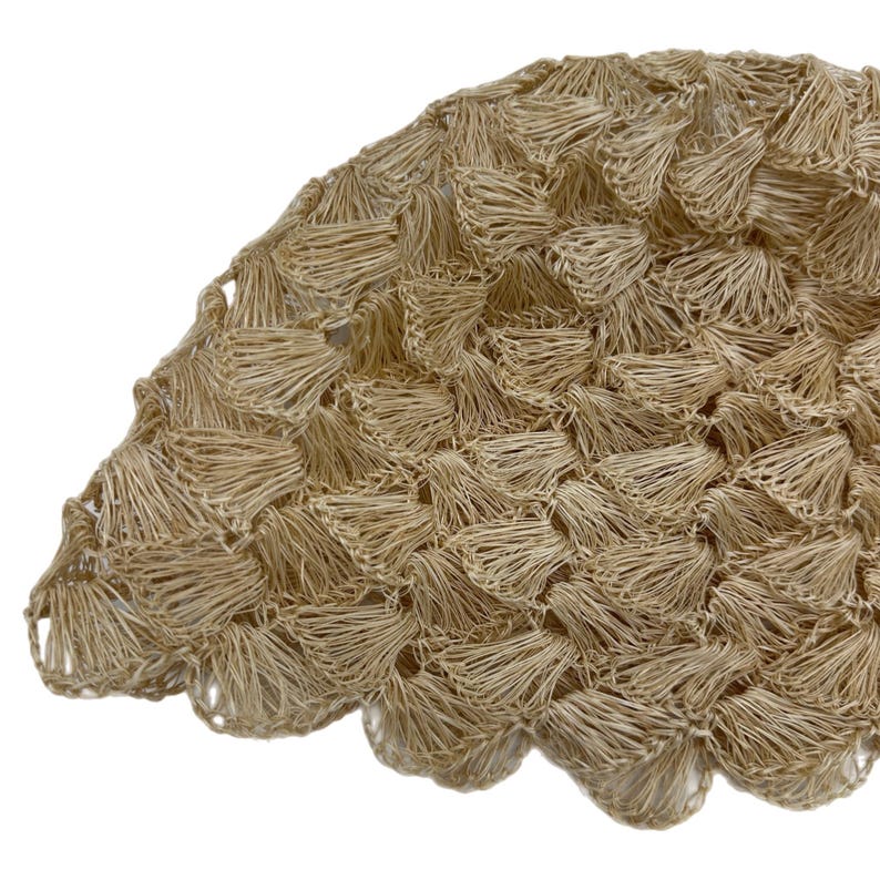 May include: Close-up of a beige, woven, circular object with a scalloped edge. The texture is dense, with a pattern of small, overlapping, tassel-like elements. The material appears to be natural fibre, possibly raffia or similar.