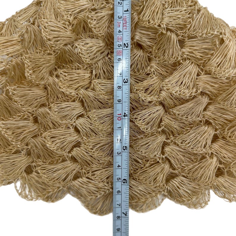May include: Close-up of a beige, textured fabric with a fringe-like design. The fabric is approximately 20 cm in length, as measured by a ruler. The material appears to be woven or crocheted, with a dense, three-dimensional texture.