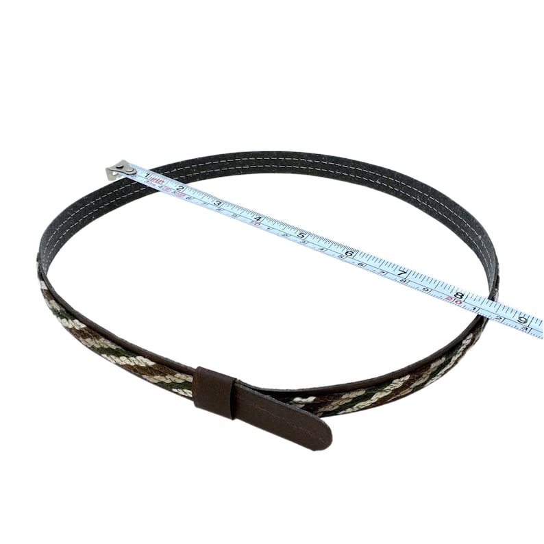 K&ouml;nnte beinhalten: Ein brauner Lederg&uuml;rtel mit einem gewebten Muster in Braun, Wei&szlig; und Gr&uuml;n. Der G&uuml;rtel wird mit einem Ma&szlig;band gemessen und ist etwa 20 cm lang.