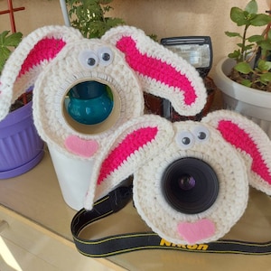 May include: Two white crocheted bunny camera lens covers with pink ears. One cover is on a camera lens and the other is on a flash.