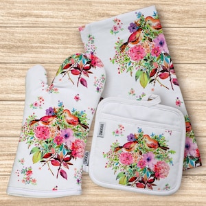 May include: A set of three kitchen items featuring a floral pattern with birds. The set includes a white oven mitt, a white pot holder, and a white tea towel. All three items have a watercolor-style floral design with pink, purple, and green flowers and two birds.
