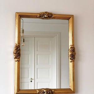 May include: A rectangular wall mirror featuring a metallic gold frame. The frame is decorated with sculpted rose motifs centered on the top, bottom, and both side edges. The mirror glass is clear and reflects the surrounding room environment.