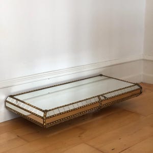 May include: A rectangular, mirrored shelf with a gold-colored metal frame. The shelf is mounted on a wall and has a clear glass surface. The frame has a decorative, geometric design. The shelf is approximately 24 inches long.