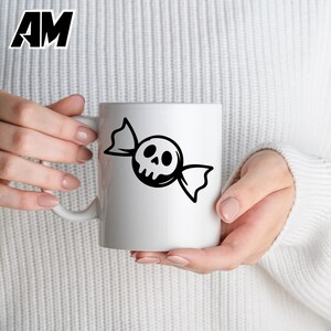 May include: White ceramic mug featuring a black candy design with a skull face. The mug has a handle and is held by a person. The letters "AM" are in the upper left corner.