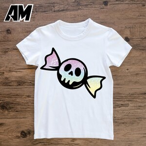 May include: White t-shirt featuring a candy-shaped skull design. The skull has a rainbow gradient, and the candy wrapper is black with pink and yellow accents. The shirt has short sleeves and a crew neck.