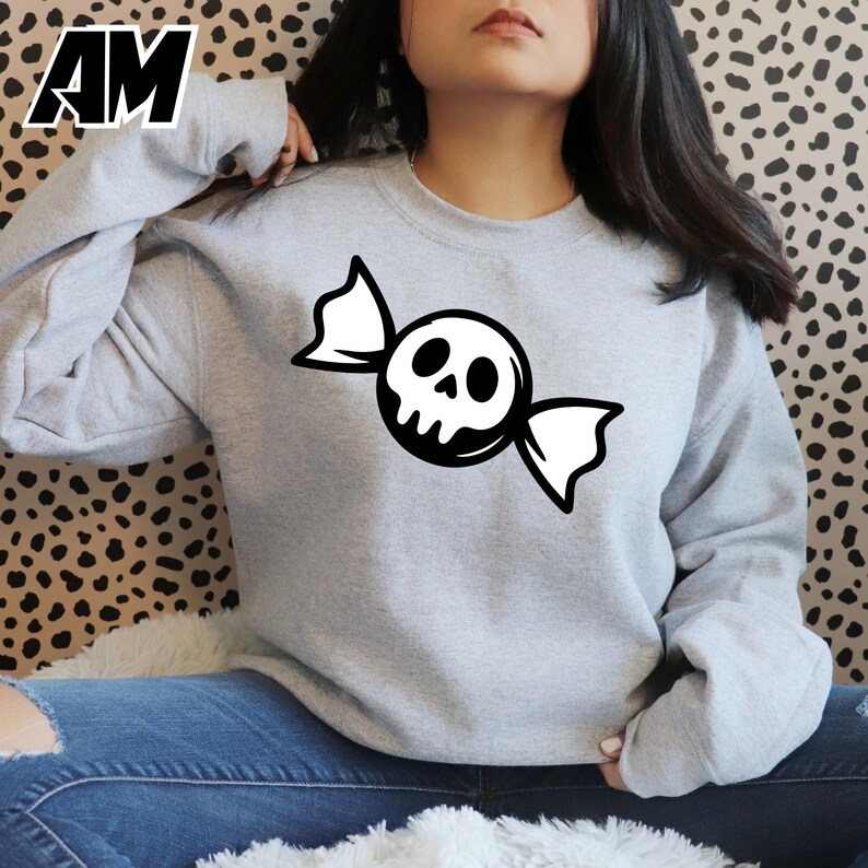 May include: A light gray sweatshirt featuring a black and white candy design with a skull face. The graphic is centered on the chest. The sweatshirt is a crew neck style.