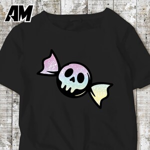 May include: Black t-shirt featuring a candy-shaped skull design. The skull is white with black eye sockets and a nose, wrapped in a colorful, glittery wrapper with pastel hues of pink, blue, and yellow. The letters "AM" are in the top left corner.