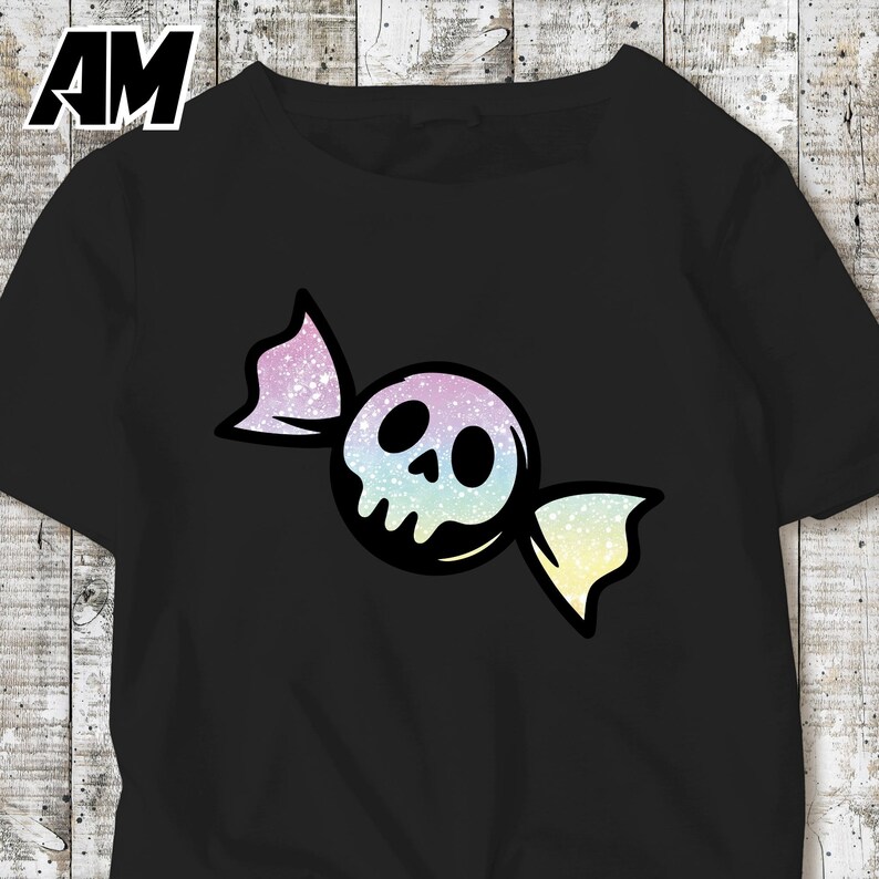 May include: Black t-shirt featuring a candy-shaped skull design. The skull is white with black eye sockets and a nose, wrapped in a colorful, glittery wrapper with pastel hues of pink, blue, and yellow. The letters "AM" are in the top left corner.