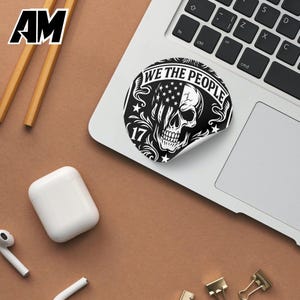 Puede incluir: Una pegatina en blanco y negro en un port&aacute;til con un dise&ntilde;o de calavera con la bandera estadounidense. La pegatina dice "WE THE PEOPLE" y "17". El port&aacute;til est&aacute; sobre una superficie marr&oacute;n, con l&aacute;pices, una funda blanca para auriculares y otros accesorios.