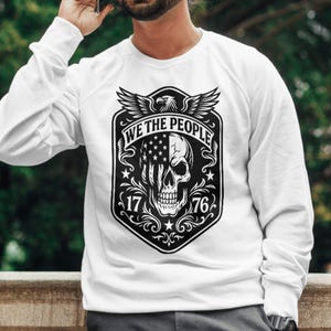 Puede incluir: Sudadera blanca con un gr&aacute;fico en blanco y negro. El gr&aacute;fico incluye una calavera con un dise&ntilde;o de la bandera estadounidense, el texto "WE THE PEOPLE", el a&ntilde;o "1776" y un emblema de &aacute;guila. La sudadera tiene mangas largas.